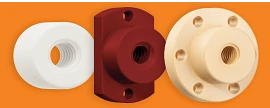 Three ACME lead screw nuts lined up left to right, the first one a white cylinder with a hole with grooves that run counterclockwise in a spiraling rotation to the back of the nut, the middle one a dark maroon red cylinder with a tall rectangle backing that has a rounded top and bottom. There are 3 holes, a small hole at the top of the rectangle and the bottom, and a larger hole with counterclockwise grooves running through it in a spiraling pattern. The nut all the way to the left is light tan/beige, a cylinder with a larger, thinner cylinder backing. The backing has 6 evenly spaced small holes around its face, and the main cylinder has a large hole with counterclockwise grooves spiraling to the back of the nut